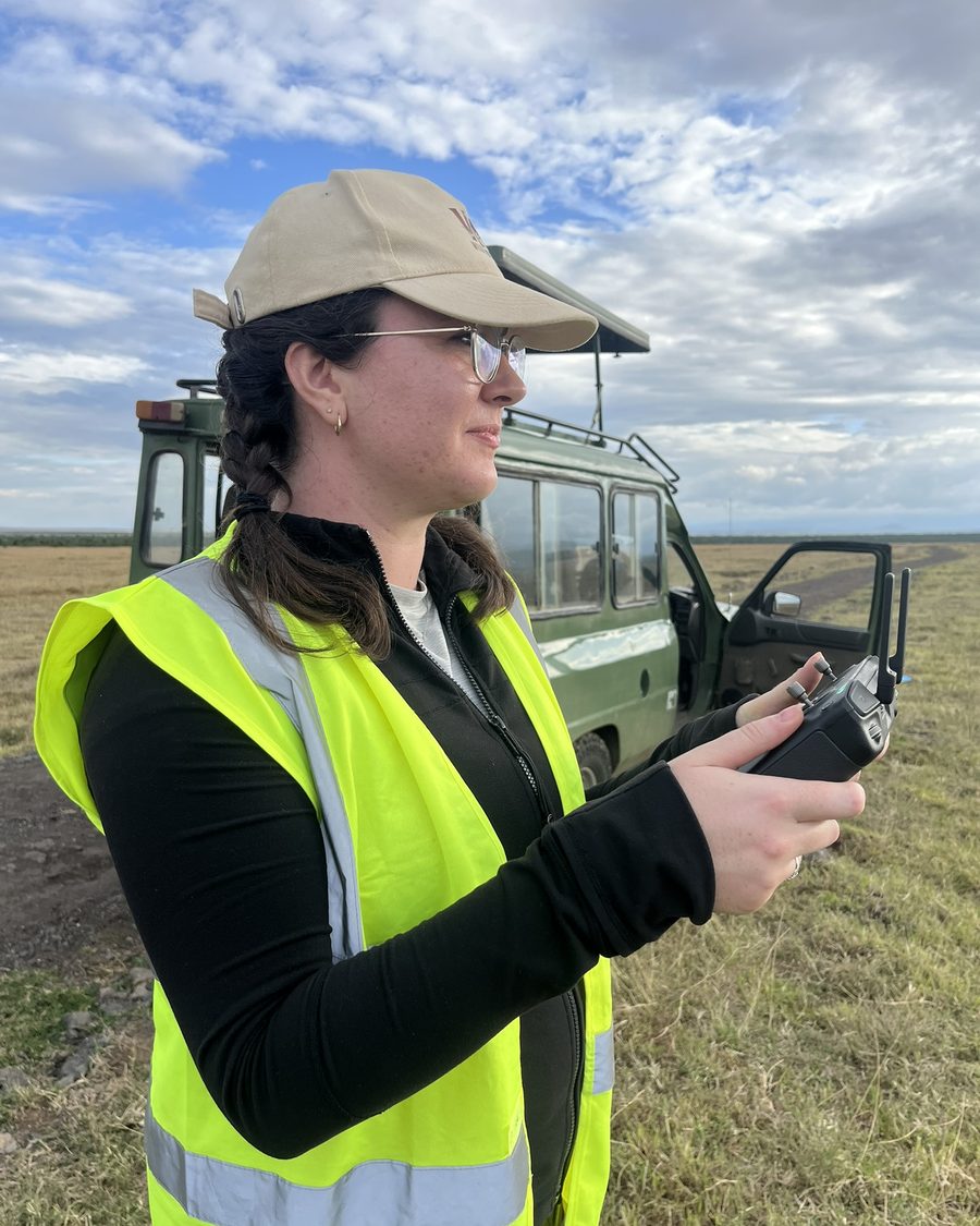 Jenna Kline piloting a drone during field research, holding a controller in an open savanna landscape.